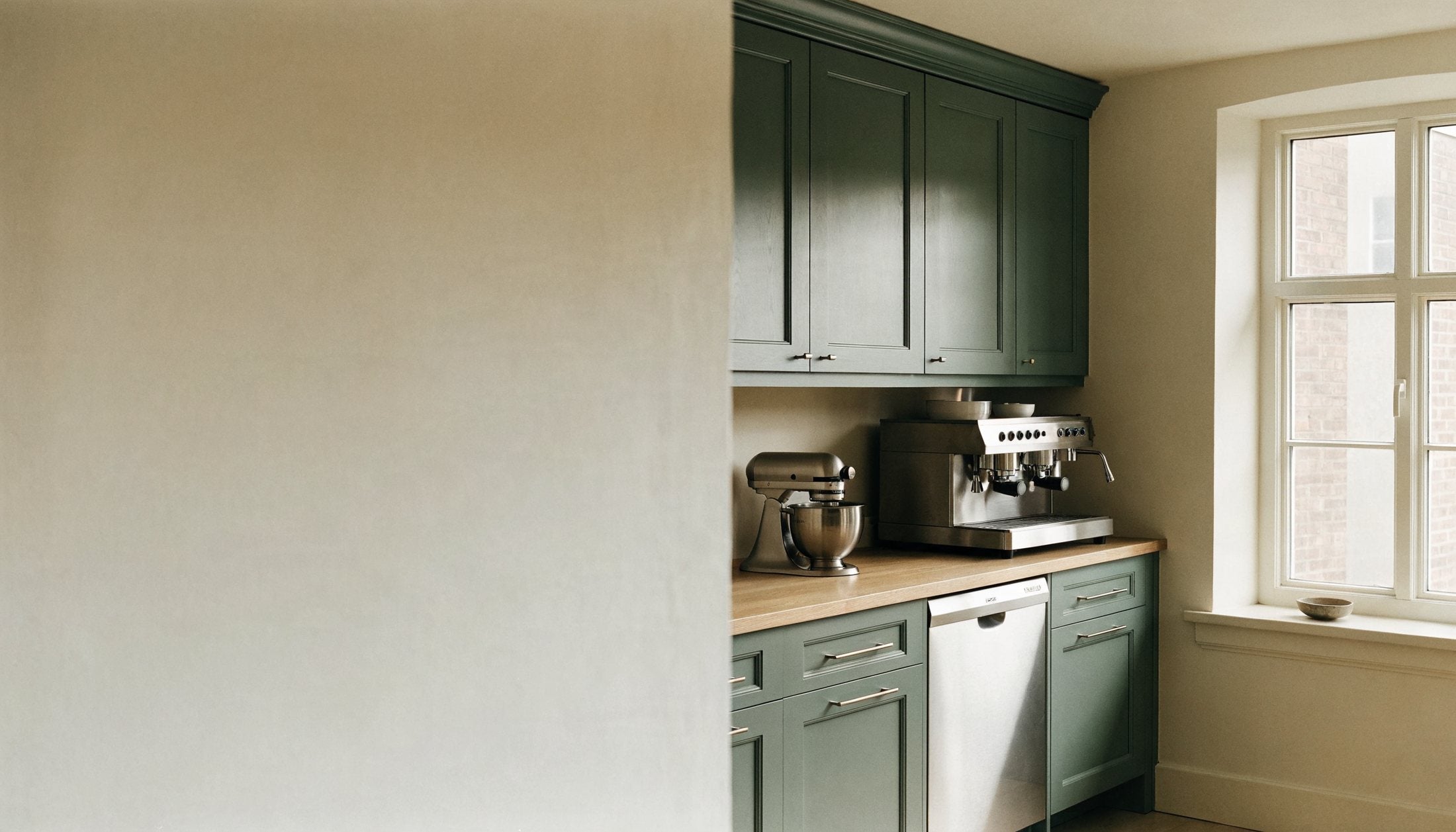 A calm kitchen in deep green cabinetry, with an espresso machine and stand mixer on an oak worktop